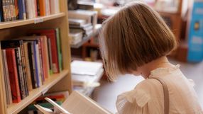 Młoda kobieta z krótkimi włosami czyta książkę w bibliotece, otoczona regałami pełnymi książek.