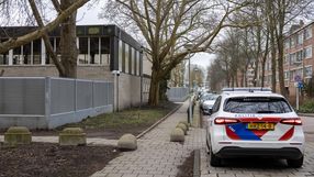 Widok na ulicę z samochodem policji, drzewami i budynkiem w tle. Pochmurne niebo.