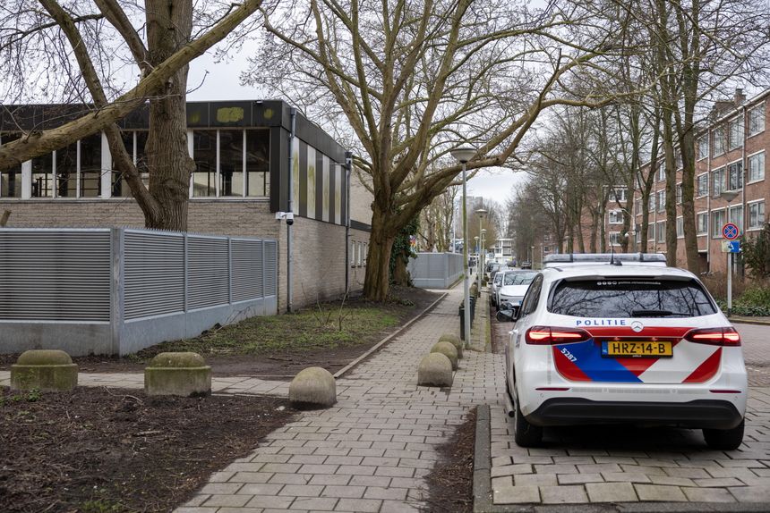 Widok na ulicę z samochodem policji, drzewami i budynkiem w tle. Pochmurne niebo.