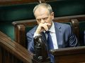 Donald Tusk w garniturze, siedzi w sejmie, z ręką na twarzy, zamyślony wśród drewnianych ław.
