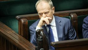 Donald Tusk w garniturze, siedzi w sejmie, z ręką na twarzy, zamyślony wśród drewnianych ław.