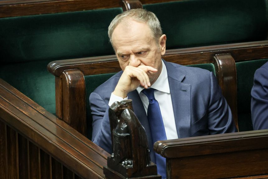 Donald Tusk w garniturze, siedzi w sejmie, z ręką na twarzy, zamyślony wśród drewnianych ław.