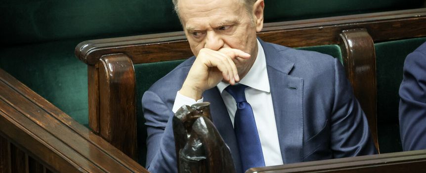 Donald Tusk w garniturze, siedzi w sejmie, z ręką na twarzy, zamyślony wśród drewnianych ław.