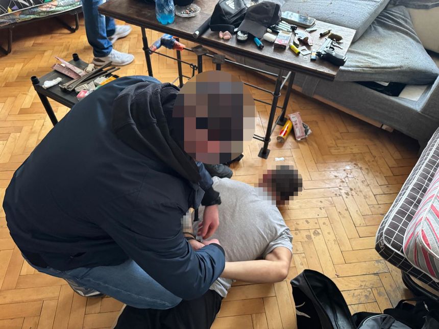 Na zdjęciu mężczyzna w szarej koszulce leży na podłodze, a inny mężczyzna go unieruchamia. W tle widać bałagan.