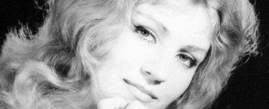 Anna German o falowanych, jasnych włosach, delikatnym uśmiechu, z dłonią przy twarzy. Czarno-białe zdjęcie.