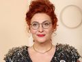 Kobieta z rudymi włosami i okularami, ubrana w bluzkę z agrafkami, uśmiecha się do kamery.