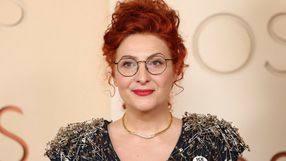 Kobieta z rudymi włosami i okularami, ubrana w bluzkę z agrafkami, uśmiecha się do kamery.