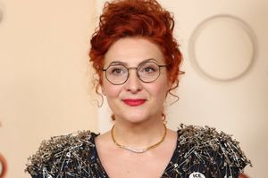 Kobieta z rudymi włosami i okularami, ubrana w bluzkę z agrafkami, uśmiecha się do kamery.