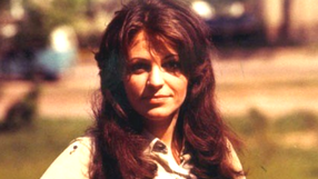 Anna Jantar z długimi, brunatnymi włosami, uśmiechająca się na tle zieleni.