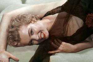 Na zdjęciu uśmiechająca się kobieta w czarnej sukience, leżąca na jasnej kanapie.