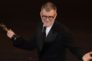 Mężczyzna w czarnym garniturze z przyznaną statuetką Oscara w ręku, na scenie.