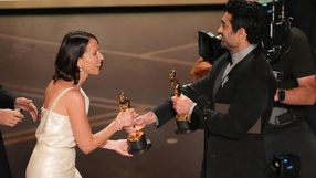 Dwie osoby w eleganckich strojach wymieniają się statuetkami oscarowymi na scenie.