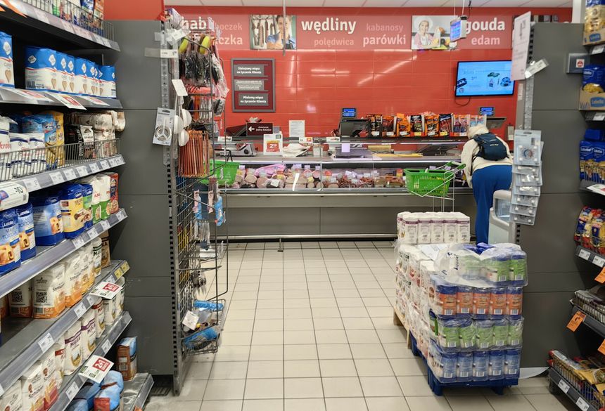 Zdjęcie przedstawia wnętrze sklepu spożywczego z regałami i ladą ze wędlinami.
