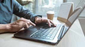 Na zdjęciu widoczna jest ręka osoby korzystającej z laptopa na biurku, obok kubek kawy.