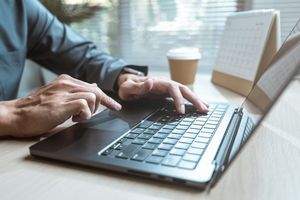 Na zdjęciu widoczna jest ręka osoby korzystającej z laptopa na biurku, obok kubek kawy.