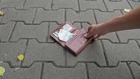 Ręka trzyma brązowy portfel leżący na chodniku, widoczne banknoty.
