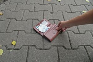 Ręka trzyma brązowy portfel leżący na chodniku, widoczne banknoty.