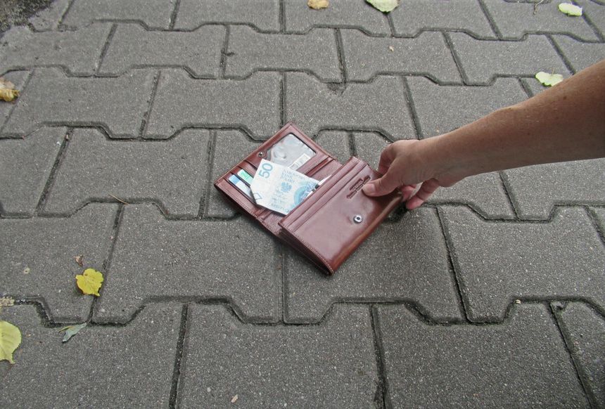 Ręka trzyma brązowy portfel leżący na chodniku, widoczne banknoty.