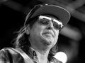 Phil Campbell w czapce i okularach przeciwsłonecznych, na tle sceny, w stroju w kratkę.