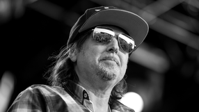 Phil Campbell w czapce i okularach przeciwsłonecznych, na tle sceny, w stroju w kratkę.