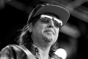 Phil Campbell w czapce i okularach przeciwsłonecznych, na tle sceny, w stroju w kratkę.