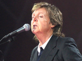 Paul McCartney z mikrofonem, ubrany w czarny garnitur, występuje na scenie. Światła w tle.