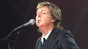 Paul McCartney z mikrofonem, ubrany w czarny garnitur, występuje na scenie. Światła w tle.
