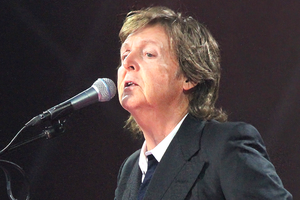 Paul McCartney z mikrofonem, ubrany w czarny garnitur, występuje na scenie. Światła w tle.