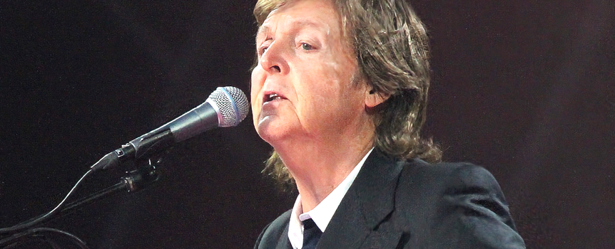 Paul McCartney z mikrofonem, ubrany w czarny garnitur, występuje na scenie. Światła w tle.