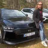 Marcin Bąkiewicz stoi obok czarnego samochodu, na tle innych aut w leśnym otoczeniu.
