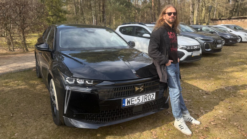 Marcin Bąkiewicz stoi obok czarnego samochodu, na tle innych aut w leśnym otoczeniu.