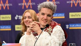 Na zdjęciu mężczyzna z jasnymi włosami, w białej bluzie, uśmiecha się do kamery. W tle kobieta.