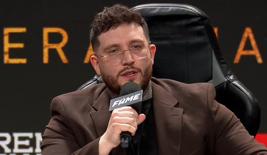 Michał „Boxdel” Baron w okularach, w ciemnej koszulce i brązowej marynarce, mówi do mikrofonu.