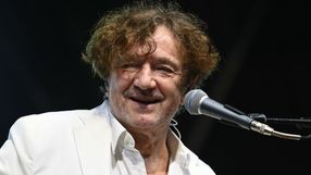 Mężczyzna z kręconymi włosami w białej marynarce, uśmiecha się do mikrofonu.