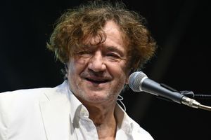 Mężczyzna z kręconymi włosami w białej marynarce, uśmiecha się do mikrofonu.