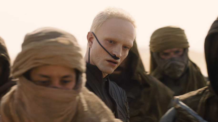Na zdjęciu grupa osób w strojach zakrywających twarze, na pierwszym planie Robert Pattinson z jasnymi włosami.