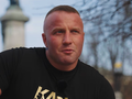 Denis Załęcki w czarnej koszulce, z tatuażem na ramieniu, siedzi w parku przy pomniku.