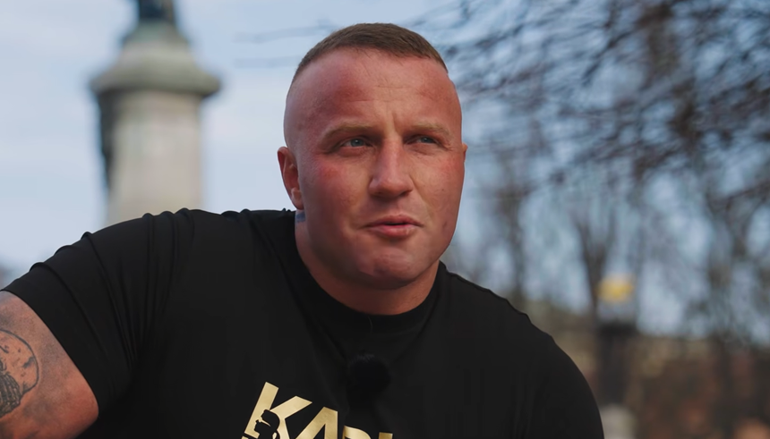 Denis Załęcki w czarnej koszulce, z tatuażem na ramieniu, siedzi w parku przy pomniku.