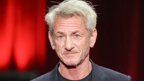 Mężczyzna w czarnym ubraniu z siwymi włosami i brodą, na tle czerwonych światł.