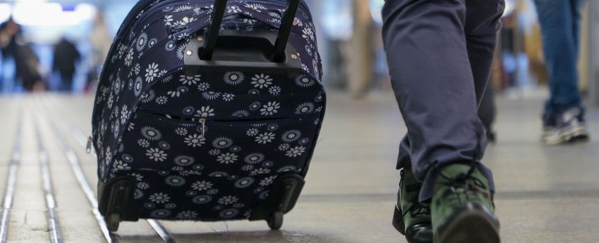 Osoba idzie korytarzem z niebieską walizką na kółkach, wokół innych podróżnych.