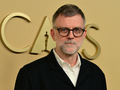 Paul Thomas Anderson w okularach, z siwą brodą, stoi przed złotym tłem z napisem 