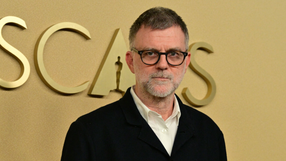 Paul Thomas Anderson w okularach, z siwą brodą, stoi przed złotym tłem z napisem 