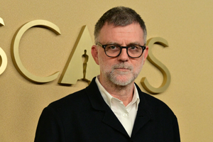 Paul Thomas Anderson w okularach, z siwą brodą, stoi przed złotym tłem z napisem 