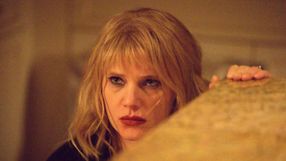 Kobieta z blond włosami patrzy intensywnie, trzymając się nieostrego przedmiotu.