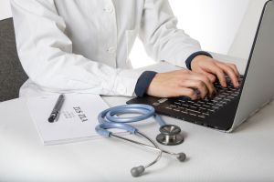 Lekarz w białym fartuchu piszący na laptopie, obok stetoskop.