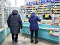 Zdjęcie przedstawia klientów apteki przy ladzie, obok pracownicy apteki. W tle regały z lekami.