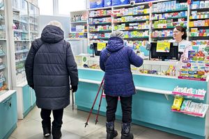 Zdjęcie przedstawia klientów apteki przy ladzie, obok pracownicy apteki. W tle regały z lekami.