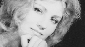 Anna German z falującymi włosami, przygląda się z uśmiechem, ręka przy twarzy. Czarno-biały portret.