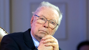 Krzysztof Zanussi w okularach, siedzi z dłońmi złożonymi, poważna mina.