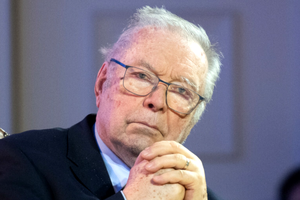 Krzysztof Zanussi w okularach, siedzi z dłońmi złożonymi, poważna mina.